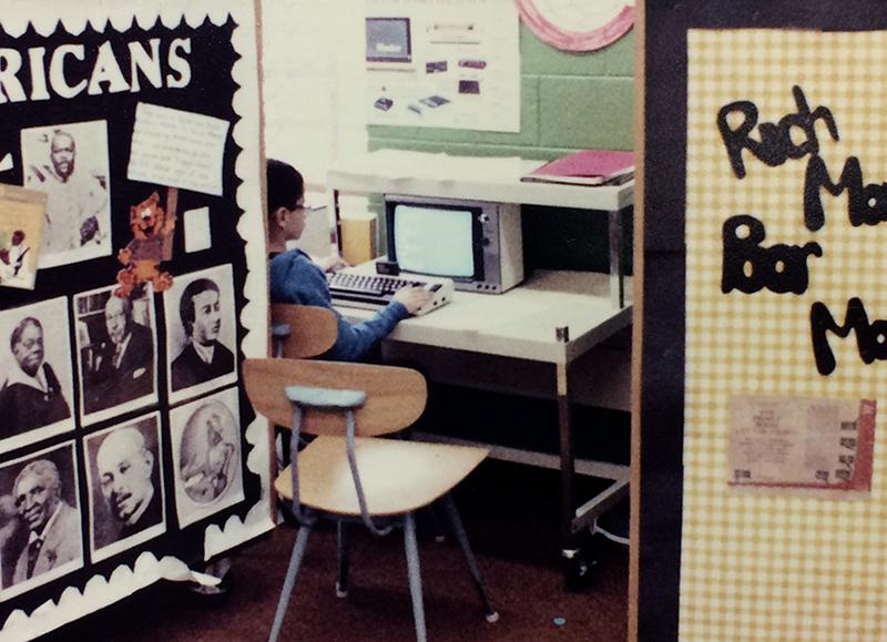 Color photograph of a student using a small personal computer. The keyboard is much thicker than keyboards today and has a cartridge slot at the top. The monitor is a small CRT screen. The student is working at a small desk behind several movable classroom storage units that also served as classroom partitions. 
