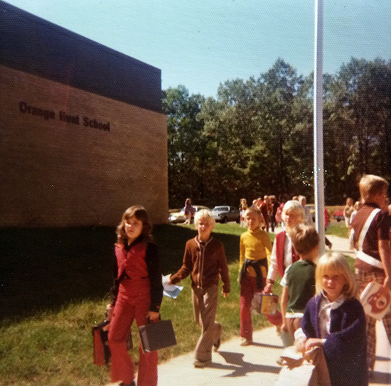 Color photograph taken on September 30, 1974. Students are walking along the sidewalk from the parking lot toward the main entrance. They are carrying backpacks and are wearing earth-toned sweaters or light jackets. The trees in the distance are still green. 