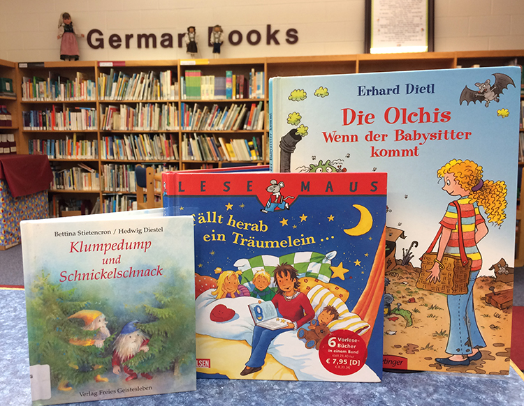 Color photograph taken in the library at Orange Hunt Elementary showing three German-language books. In the distance the German Books section of the library can be seen. The shelves hold hundreds of books. 