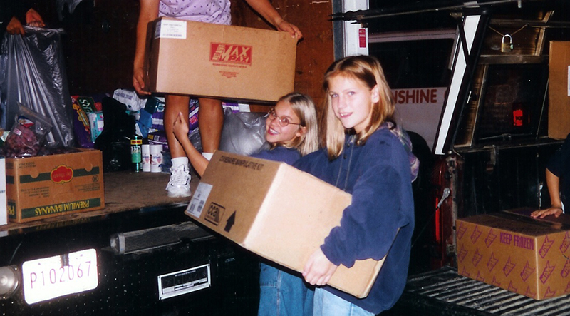 Color photograph of two students who are assisting with loading boxes into the back of a U-haul-style truck as part of a hurricane relief effort. Some of the boxes contain food, clothing, household supplies, and cleaning products. 