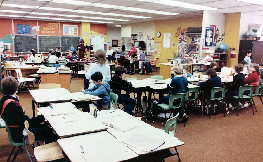 Color photograph of one of the open classrooms at Orange Hunt Elementary School. Individual desks have been clustered together in several groupings. The students in the foreground are working on one lesson, and in the distance a separate group of students are working on a different lesson.