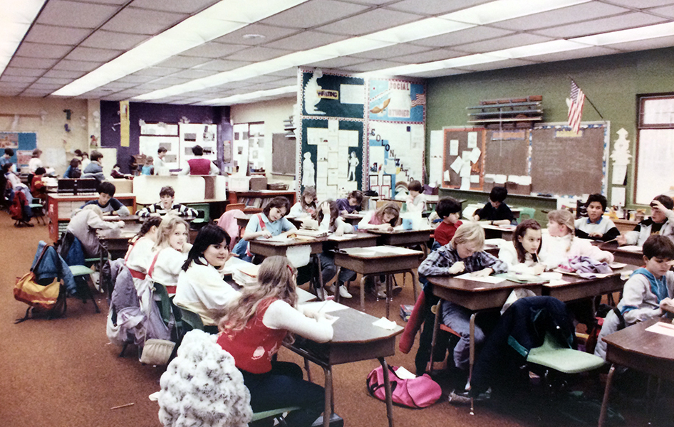 Color photograph of one of the open classrooms at Orange Hunt Elementary School. Individual desks have been clustered together in several, mostly linear, groupings of three or four children. The students in the foreground are working on one lesson, and in the distance a separate group of students can be seen working on a different lesson with another teacher. 