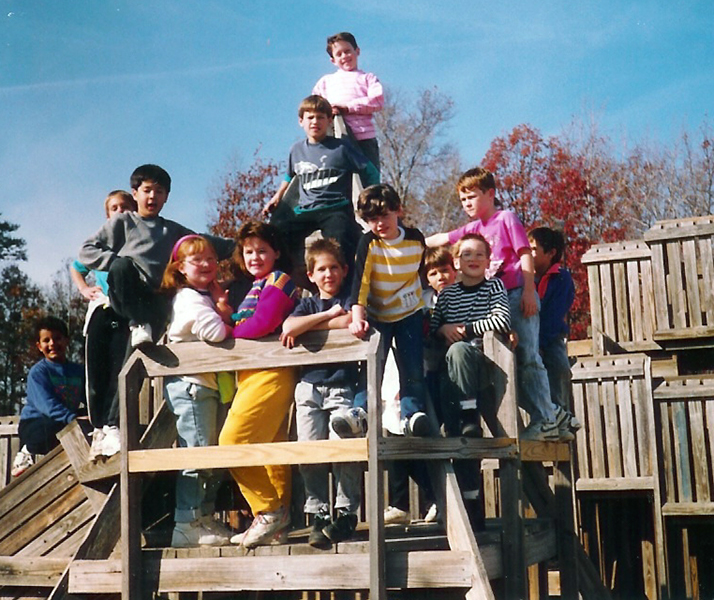 Color photograph of students posing on the wooden playground built in 1983. It is a crisp fall day. In the distance some trees have lost their leaves and others are bright red in color. 