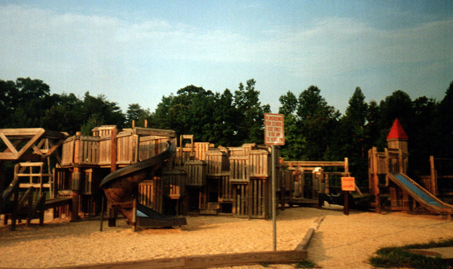 Color photograph of the playground that was built in 1983. It is a wooden structure with slides, ramps, turrets, and climbing structures. 