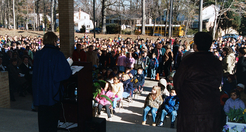 Color photograph taken during the re-dedication ceremony. The photographer is standing near the front doors of our school looking out toward the bus loop. Principal Barbee can be seen standing at a podium addressing the entire student body and a large group of parents and community members who are gathered outside in front of the building to watch the ribbon cutting ceremony. 