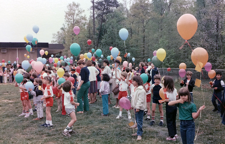 Color photograph of a large group of students and teachers outside Orange Hunt Elementary School on the playground field. The children hold colorful balloons that have small paper notes affixed to them by string. The children are waiting for the signal to release the balloons. 