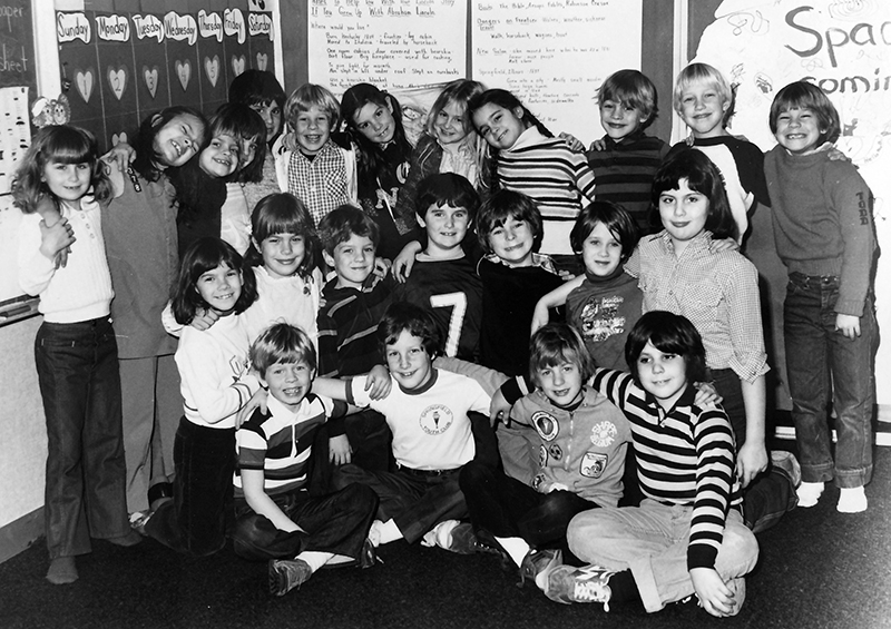 Black and white photograph of a large group of students posing for the camera. From the look of their clothing, the picture was likely taken during the late 1970s or early 1980s. 23 students are pictured, 10 of whom are girls. All the children are hugging and smiling, with their arms around one another. The photograph was taken in a classroom and there are handwritten lesson notes and a calendar on the wall behind them. 