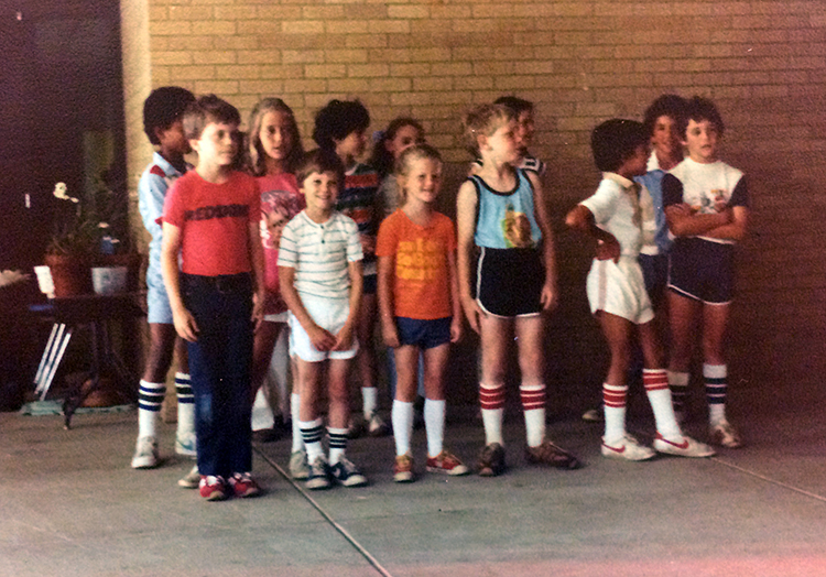 A group of 12 students of multiple ages, boys and girls, are standing next to the brick wall on the outside of our building. Some appear to be posing for another camera and others are engaged in conversation. 