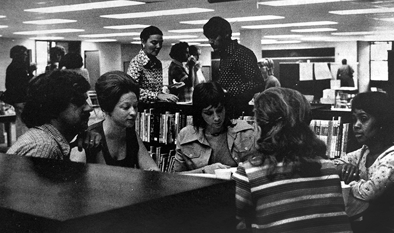 Black and white photograph of several groups of teachers working together in the library at Lake Braddock Secondary School. In the foreground, a group of five teachers are seated close to one another and appear to be engaged in a discussion of some sort. The picture appeared in the November 1974 FCPS Bulletin publication.