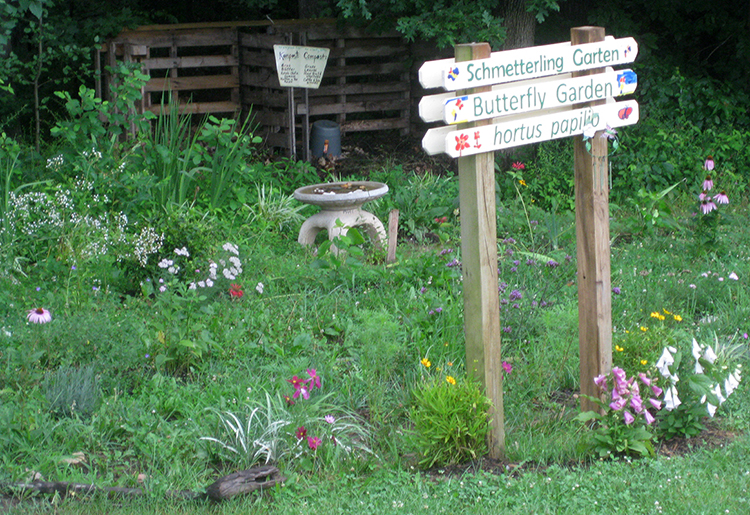 Photograph of the butterfly garden at Orange Hunt Elementary School. The garden plants are lush and green and many different types of flowers are visible. A hand-painted sign greets visitors to the garden. It has the words Butterfly Garden written on it in both English and German. A compost bin is visible in the far background. 